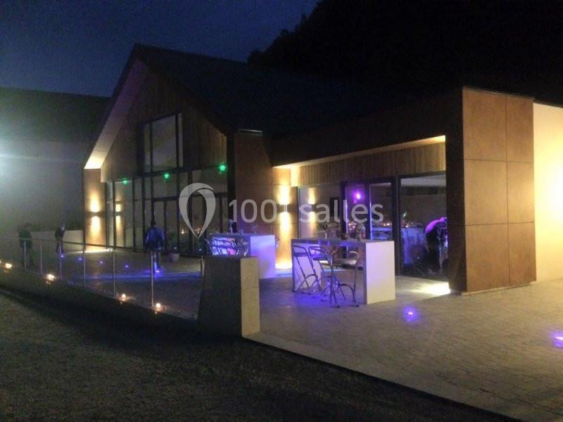 Bâtiment moderne éclairé de nuit avec de grandes baies vitrées et une terrasse aménagée.