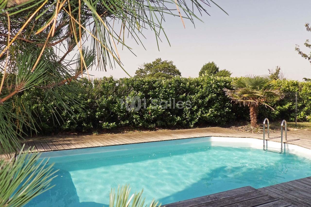 Piscine extérieure entourée de végétation, avec une terrasse en bois et un ciel dégagé en arrière-plan.