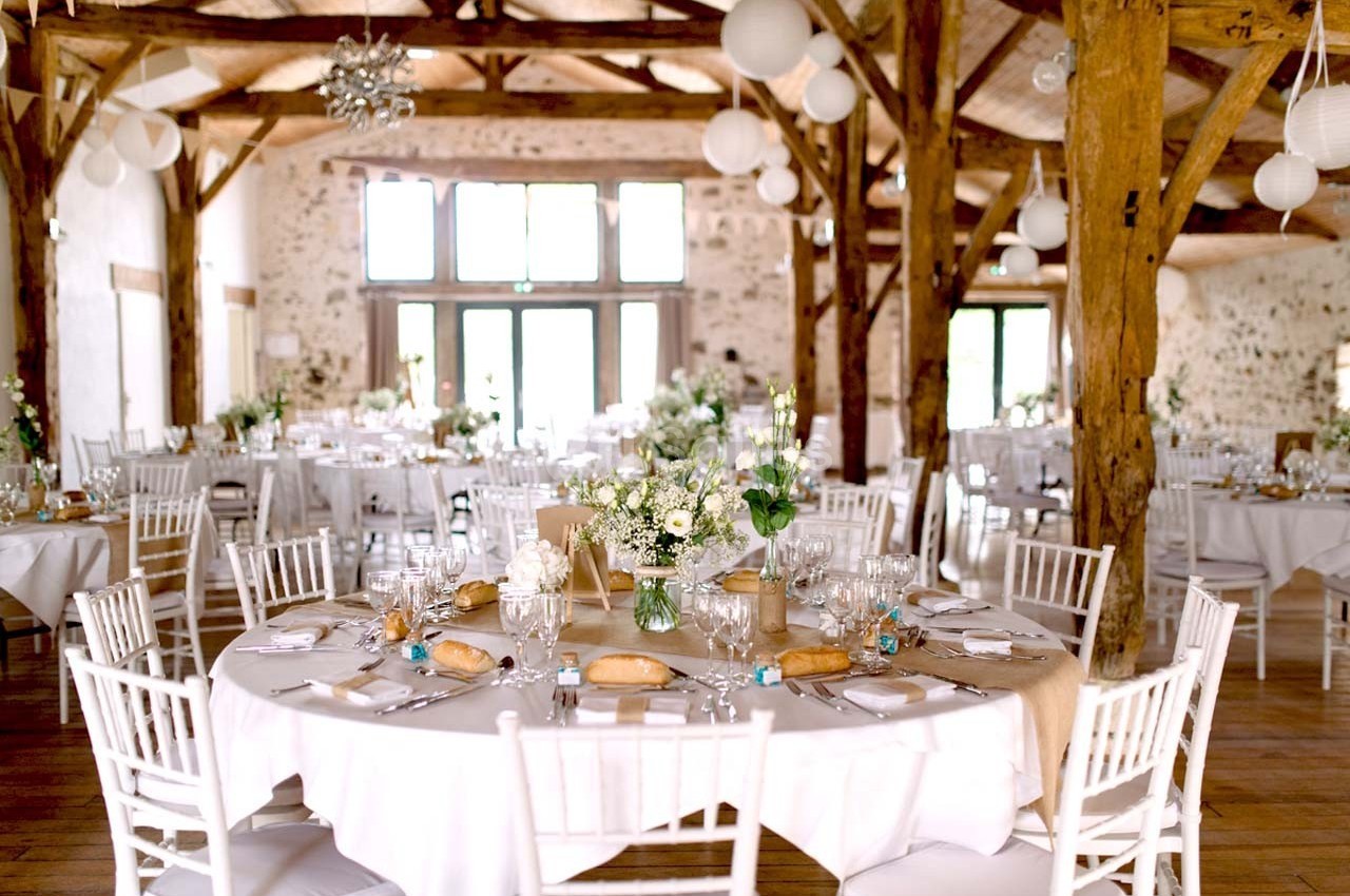 Salle de réception lumineuse avec tables rondes décorées de fleurs blanches, chaises blanches et poutres en bois apparentes.