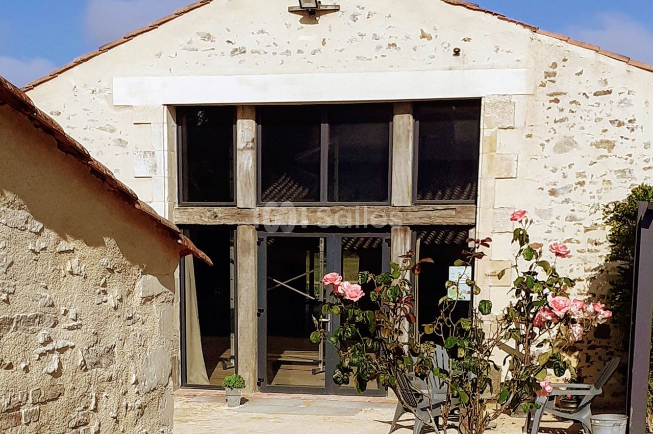 Façade d'un bâtiment en pierre avec de grandes baies vitrées, entouré d'une cour et d'un rosier en fleurs au premier plan.