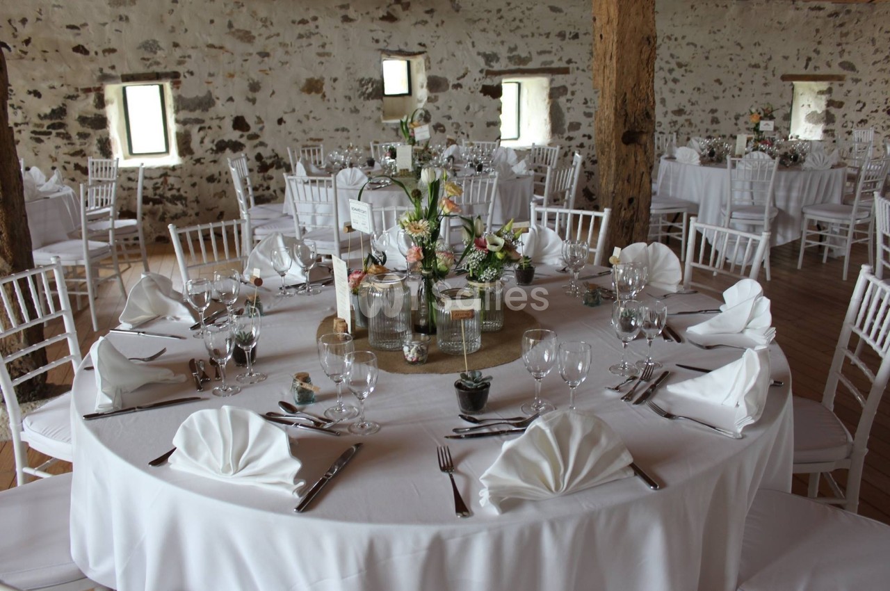Salle de réception avec tables rondes dressées, nappes blanches, serviettes pliées et décoration florale centrale.
