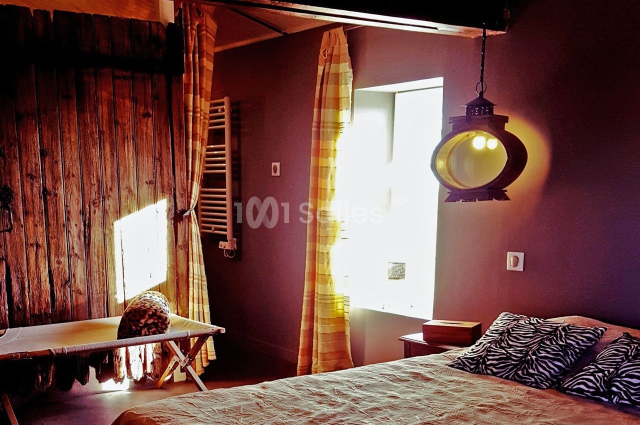 Chambre rustique avec lit, lumière naturelle entrant par une fenêtre, murs en bois et lampe suspendue.