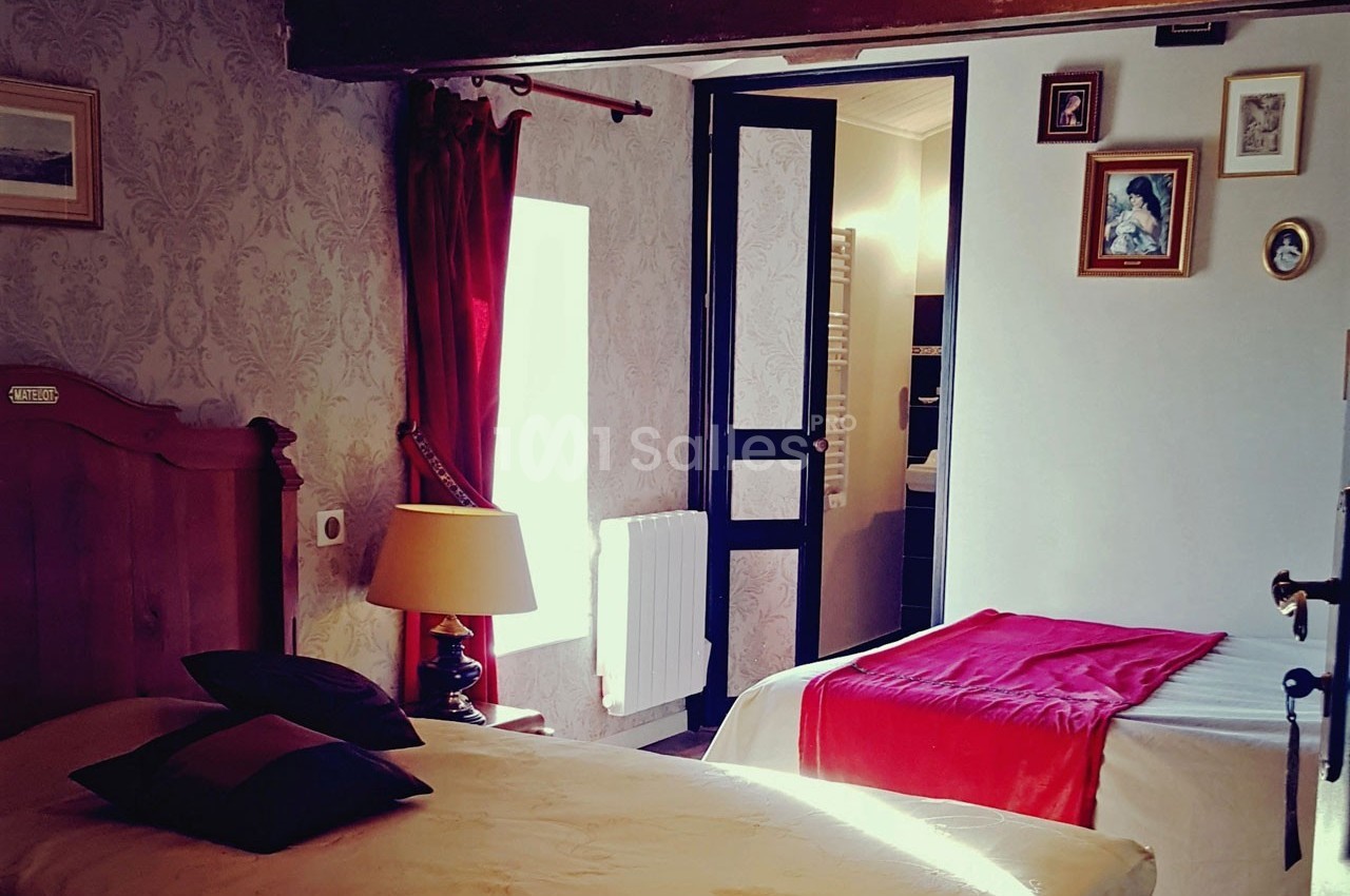 Chambre avec deux lits, murs décorés de cadres, rideaux rouges et lumière naturelle entrant par une fenêtre.