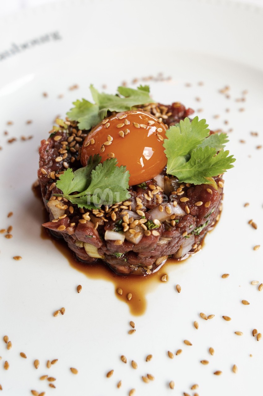 Tartare de bœuf garni de coriandre, jaune d'œuf et graines de sésame, présenté sur une assiette blanche.