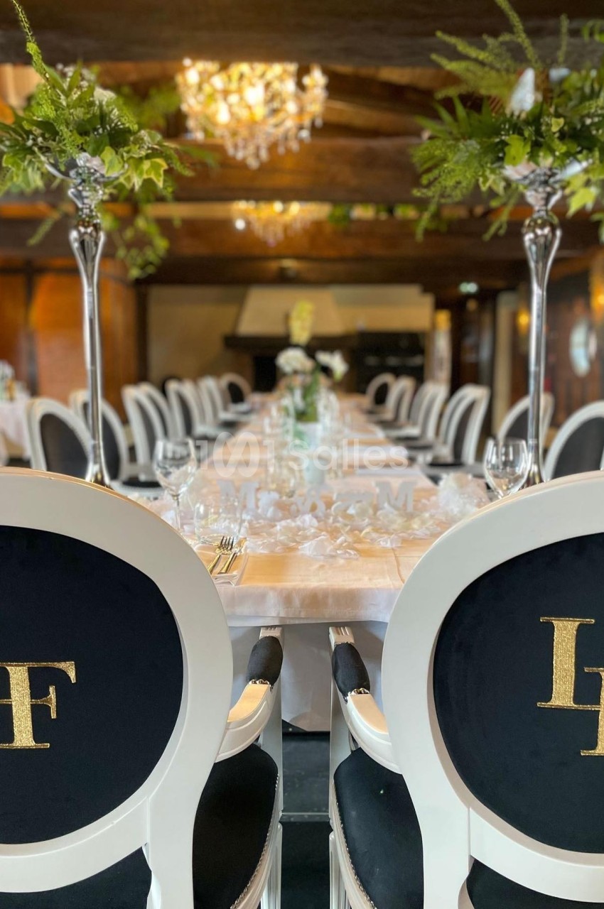 Salle de réception élégante avec tables dressées, chaises noires et blanches, chandeliers et décorations florales.