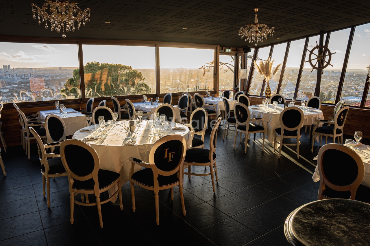 Salle de restaurant élégante avec grandes baies vitrées offrant une vue panoramique sur une ville au coucher du soleil.
