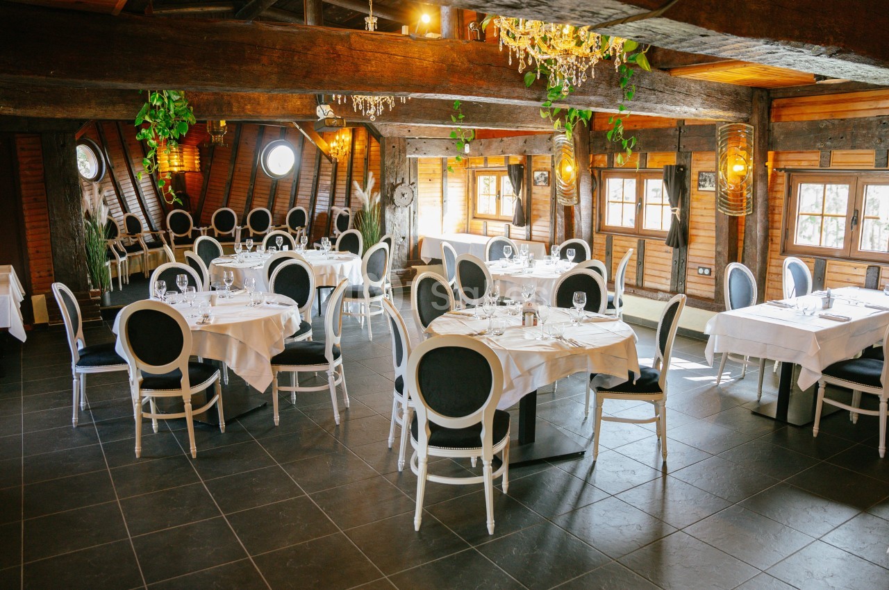 Salle de restaurant lumineuse avec tables dressées, chaises élégantes et poutres apparentes en bois.