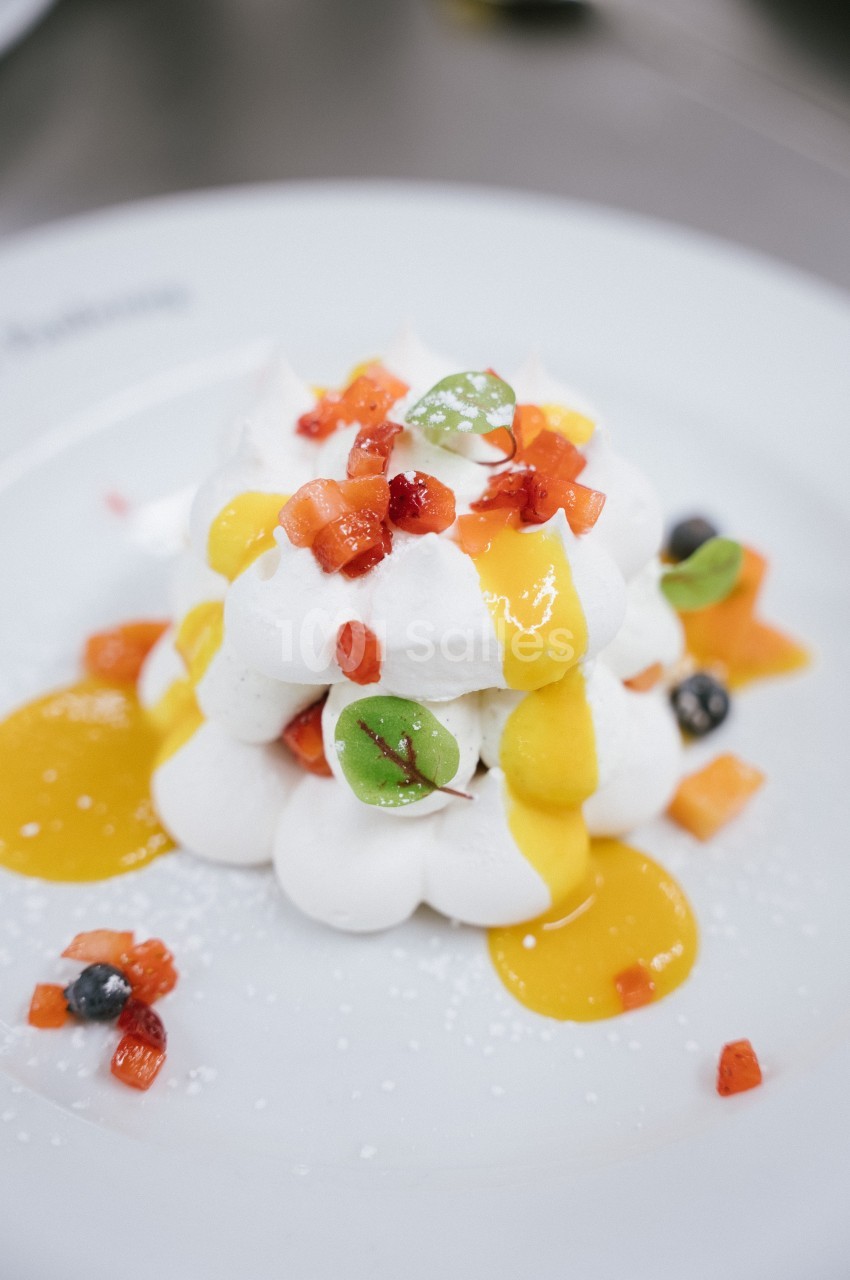 Dessert composé de meringue, coulis de mangue, morceaux de fruits rouges et feuilles décoratives sur une assiette blanche.