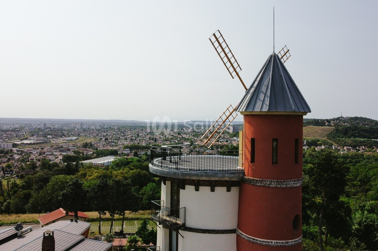 Vue d'un moulin à vent rouge avec toit en ardoise, surplombant une ville et des collines verdoyantes en arrière-plan.