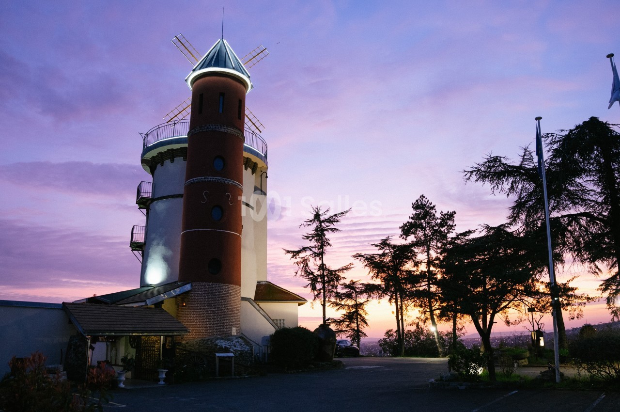 Tour en briques rouges avec des moulins à vent au sommet, éclairée au crépuscule, entourée d'arbres et d'un ciel violet.