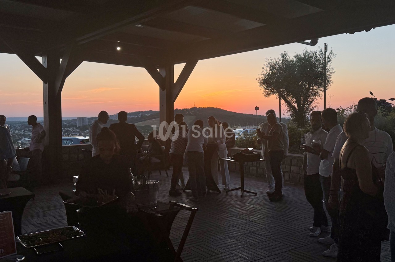 Groupe de personnes réunies sous une terrasse ouverte au coucher du soleil, avec vue sur un paysage vallonné.