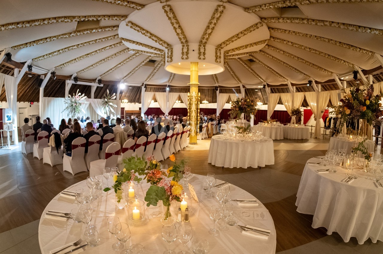 Salle de réception décorée avec des tables dressées, des chaises alignées et des fleurs, prête pour un événement.