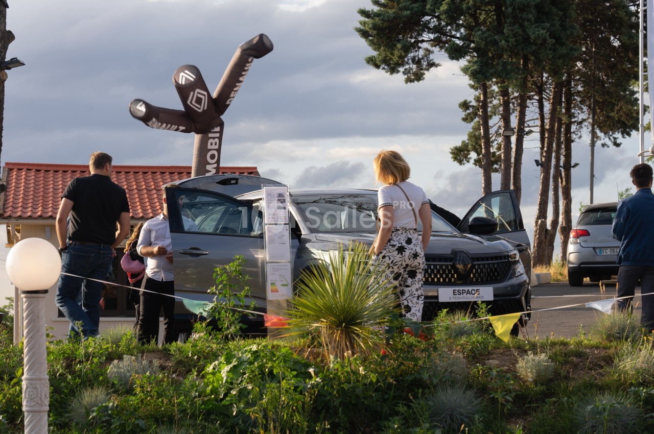 Des personnes observent une voiture exposée en extérieur, entourée de plantes et d'un bonhomme gonflable publicitaire.
