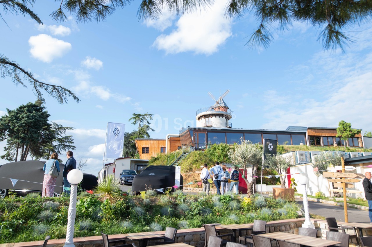 Vue d'un bâtiment moderne avec un moulin, entouré de végétation et de personnes en extérieur par temps ensoleillé.