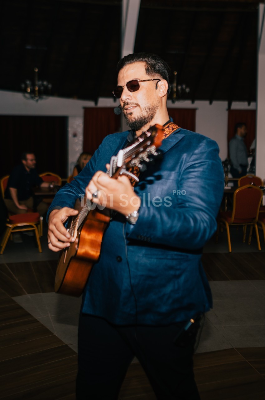 Un homme en veste bleue joue de la guitare dans une salle avec des tables et des chaises rouges en arrière-plan.