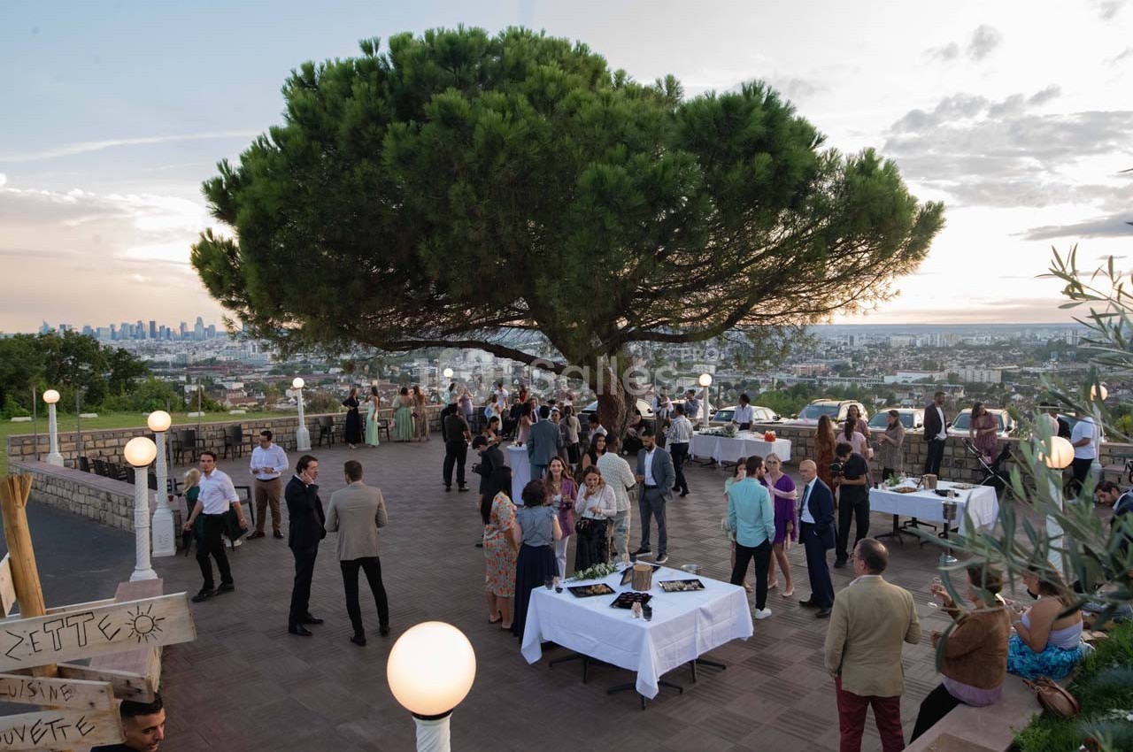 Groupe de personnes rassemblées sur une terrasse avec vue panoramique, autour de tables, sous un grand pin au coucher du…