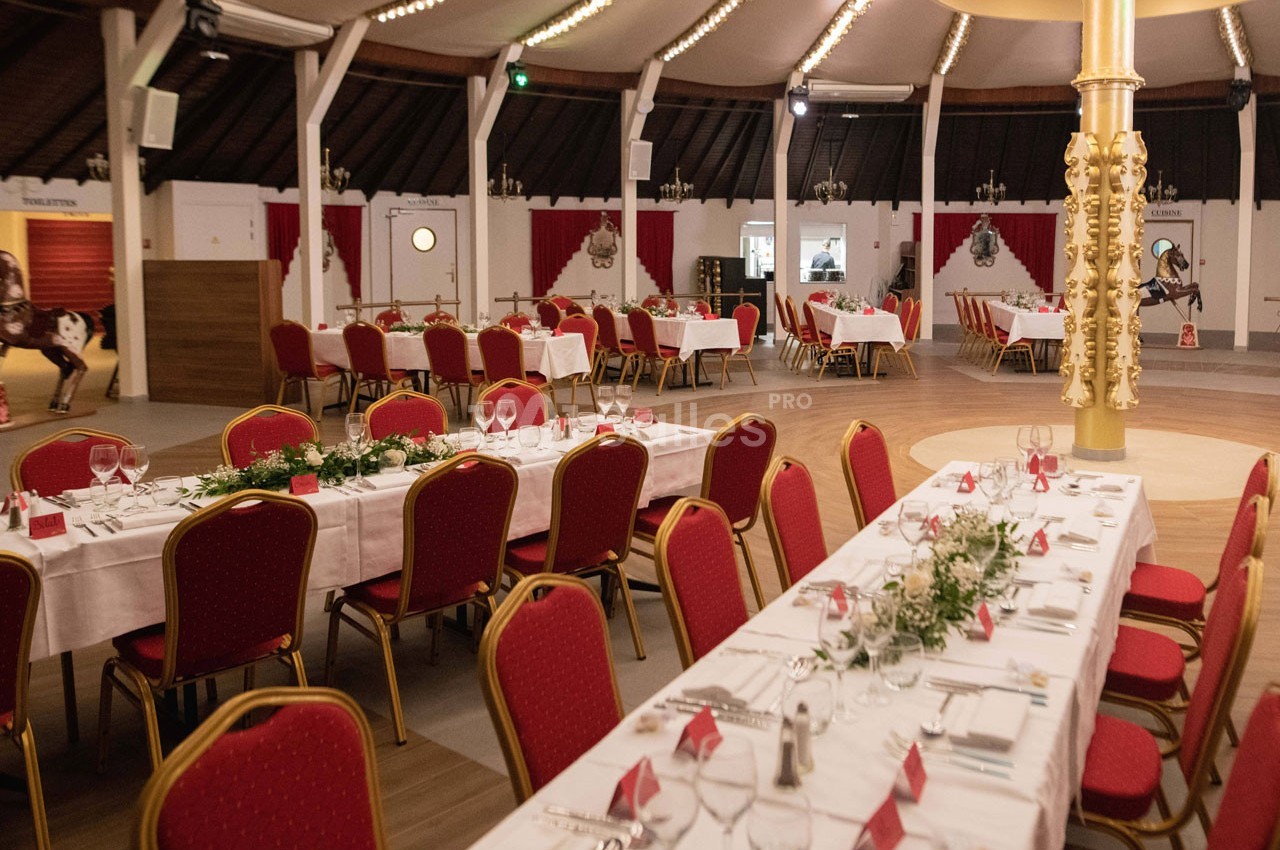 Salle de réception décorée avec des tables dressées, nappes blanches, chaises rouges et ornements végétaux.
