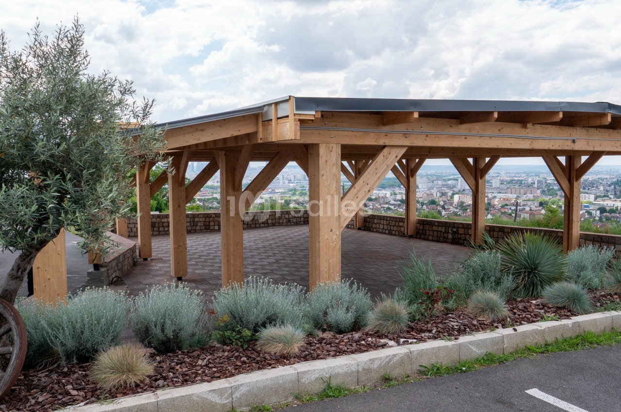 Structure en bois ouverte avec toit, entourée de plantes et offrant une vue panoramique sur une ville en arrière-plan.