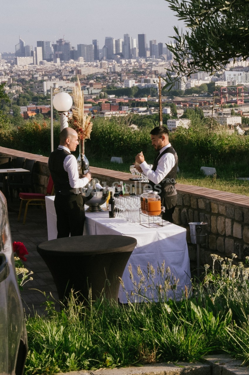 Deux serveurs en tenue formelle préparent une table avec boissons et verres, en extérieur avec vue sur une ville.