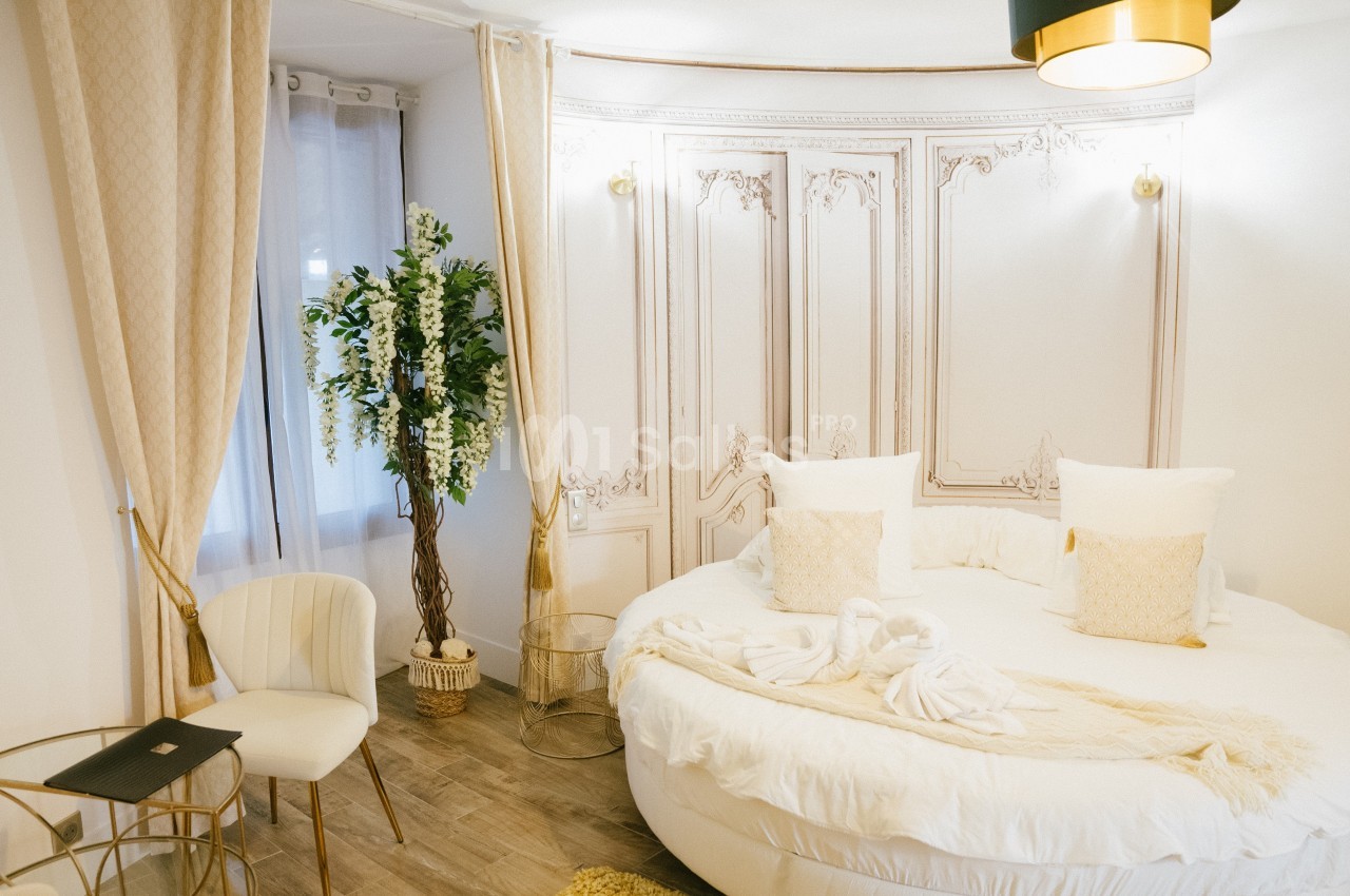 Chambre lumineuse avec lit rond, décor élégant, rideaux beiges, plante verte et mobilier doré sur parquet clair.
