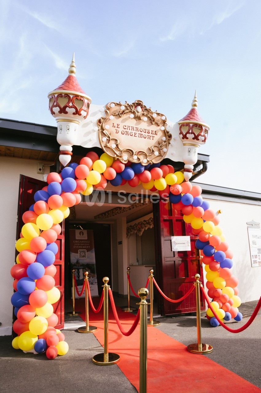 Entrée décorée avec une arche de ballons colorés et un tapis rouge menant à un bâtiment au style festif.