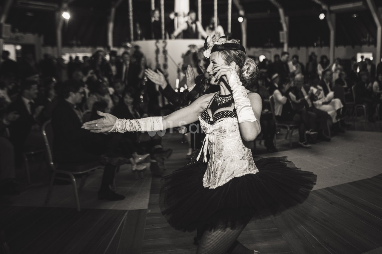 Danseuse en tenue de cabaret tenant un micro, performe devant un public assis dans une salle sombre.