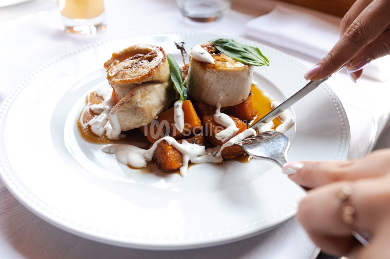Personne coupant un plat composé de médaillons de viande, légumes rôtis et sauce blanche, servi sur une assiette blanche.