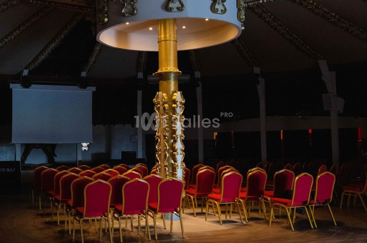 Chaises rouges disposées en cercle autour d'une colonne dorée dans une salle sombre avec un éclairage central.