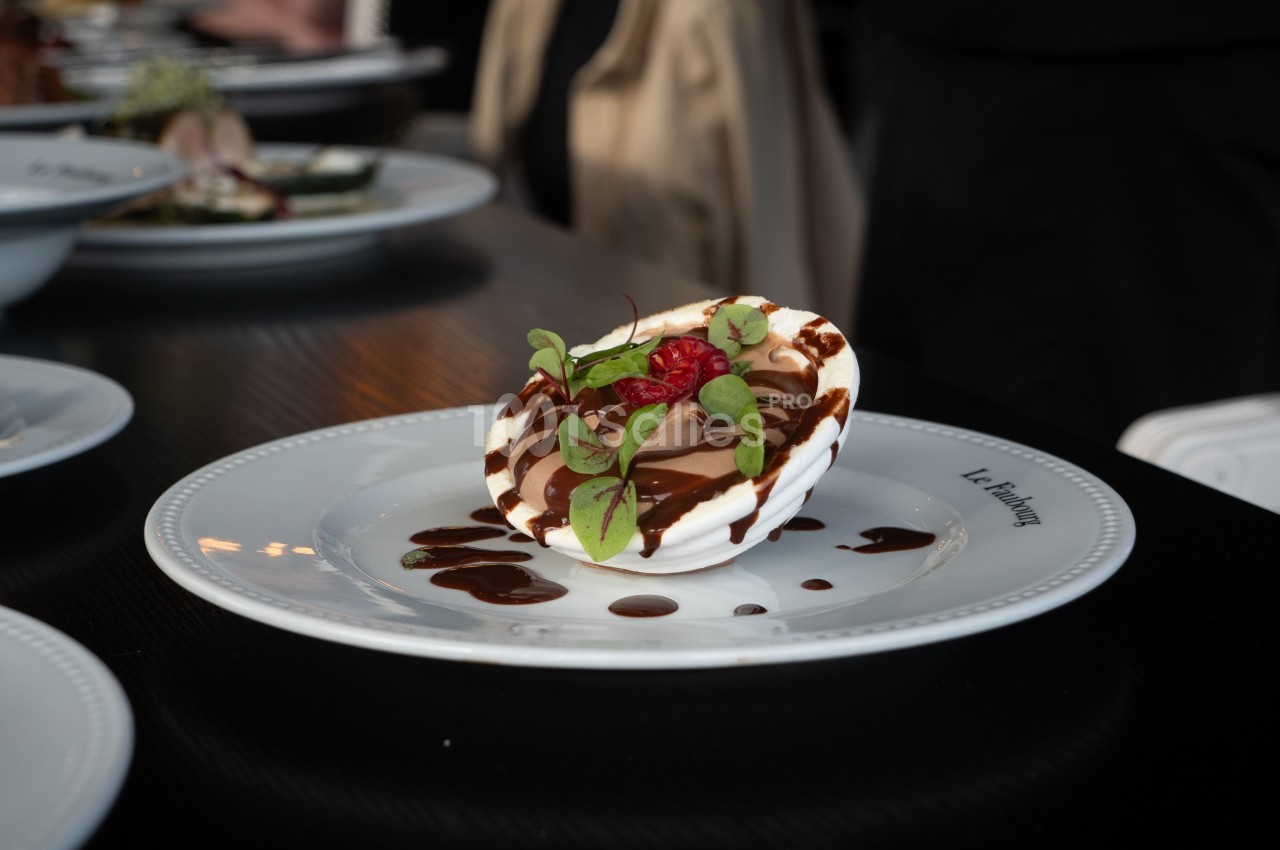Dessert dressé sur une assiette blanche, garni de crème, chocolat, framboise et feuilles vertes, avec sauce chocolatée.