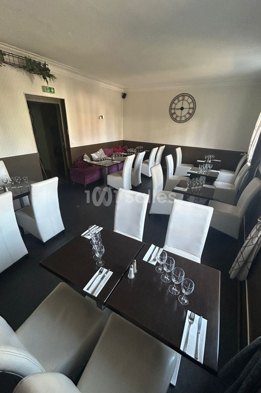 Salle de restaurant avec tables dressées, chaises blanches et décoration sobre, éclairée par la lumière naturelle.