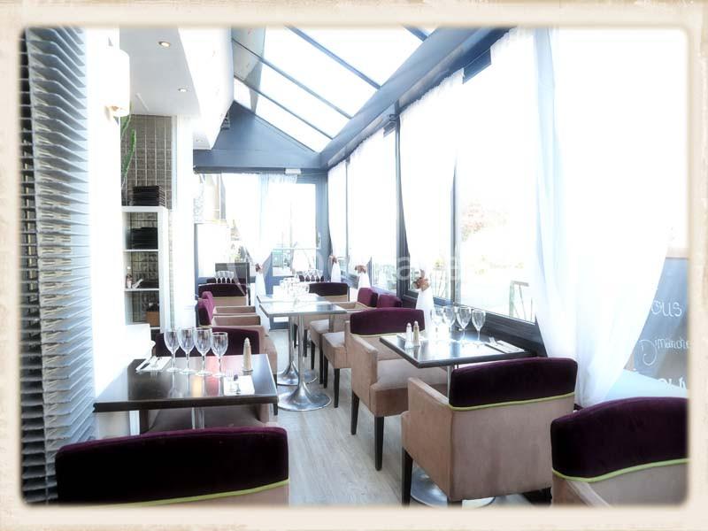 Salle de restaurant lumineuse avec de grandes baies vitrées, tables dressées et chaises rembourrées.