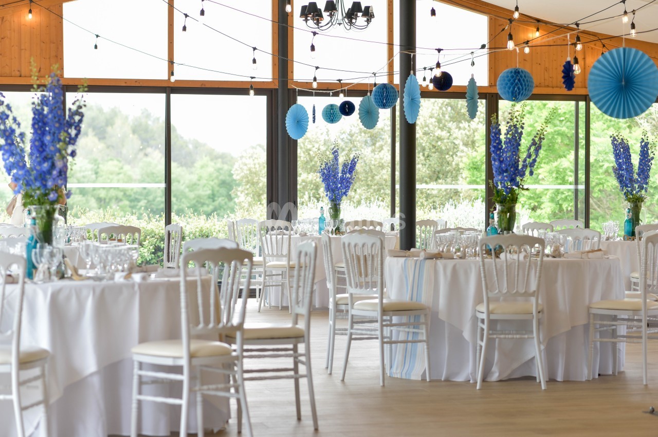 Salle de réception lumineuse décorée de guirlandes, fleurs bleues et tables rondes dressées avec nappes blanches.