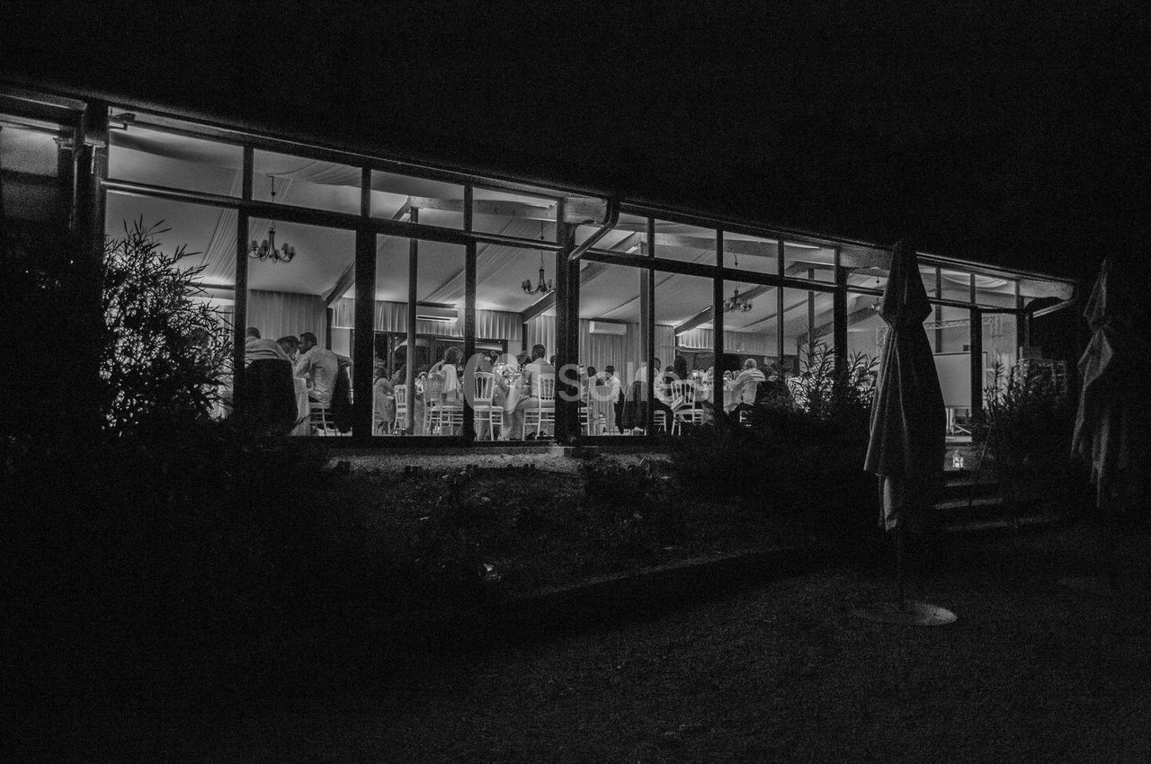 Vue nocturne d'une salle vitrée éclairée où des personnes sont réunies autour de tables.
