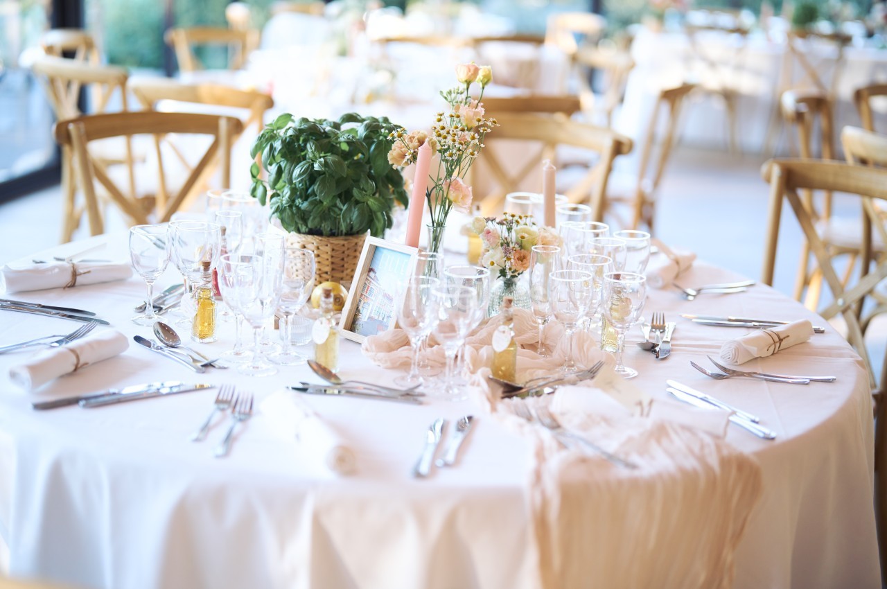 Table élégamment dressée avec nappes blanches, verres, couverts, décorations florales et plante en pot au centre.