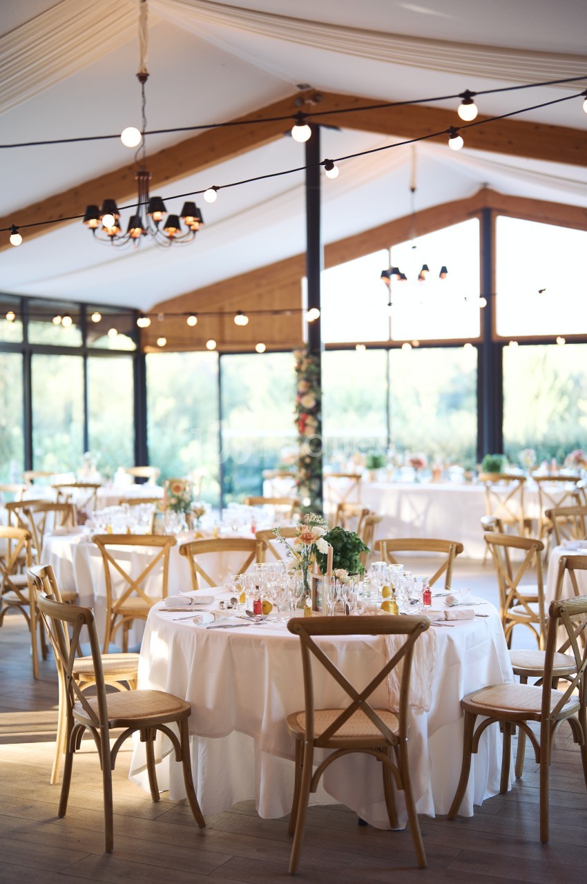 Salle de réception lumineuse avec tables rondes décorées, chaises en bois et grandes baies vitrées donnant sur un extérieur…