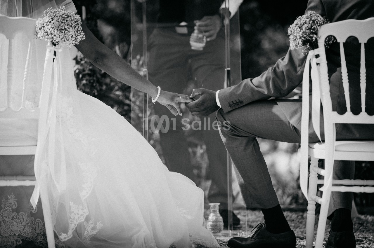 Un couple assis se tenant la main lors d'une cérémonie de mariage en extérieur, capturé en noir et blanc.