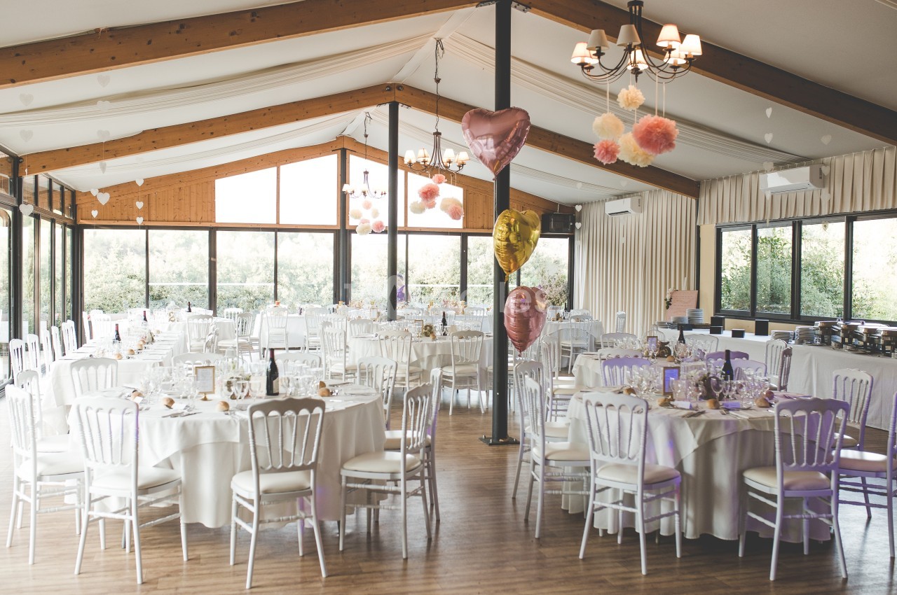 Salle de réception lumineuse décorée pour un événement, avec tables rondes, chaises blanches et ballons colorés.