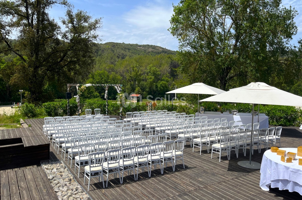 Chaises blanches alignées en extérieur sur une terrasse en bois, avec une arche décorée et des parasols sous un ciel dégagé.