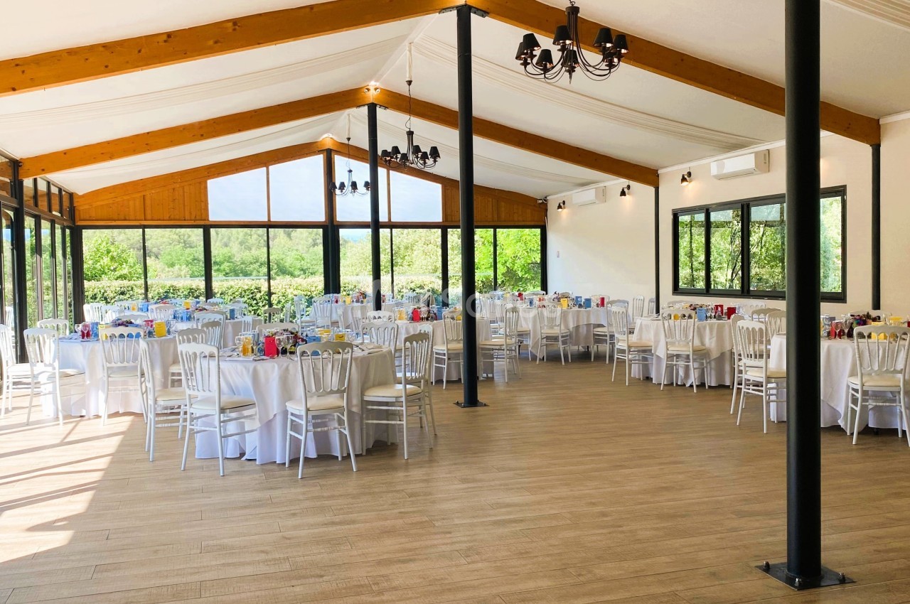 Salle lumineuse avec de grandes baies vitrées, tables rondes dressées avec nappes blanches et chaises alignées.