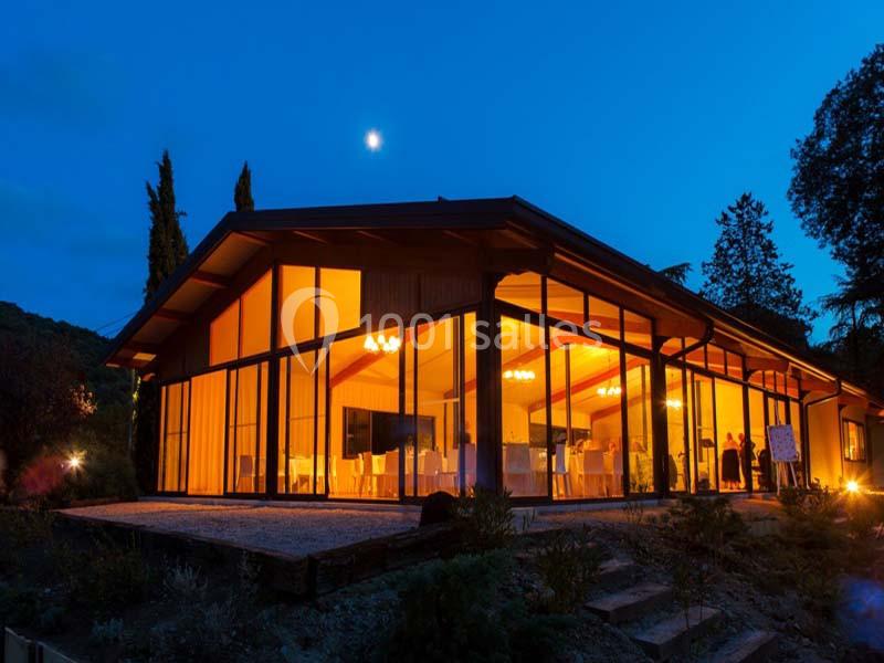 Vue nocturne d'un bâtiment vitré illuminé, entouré de végétation et éclairé par la lune.