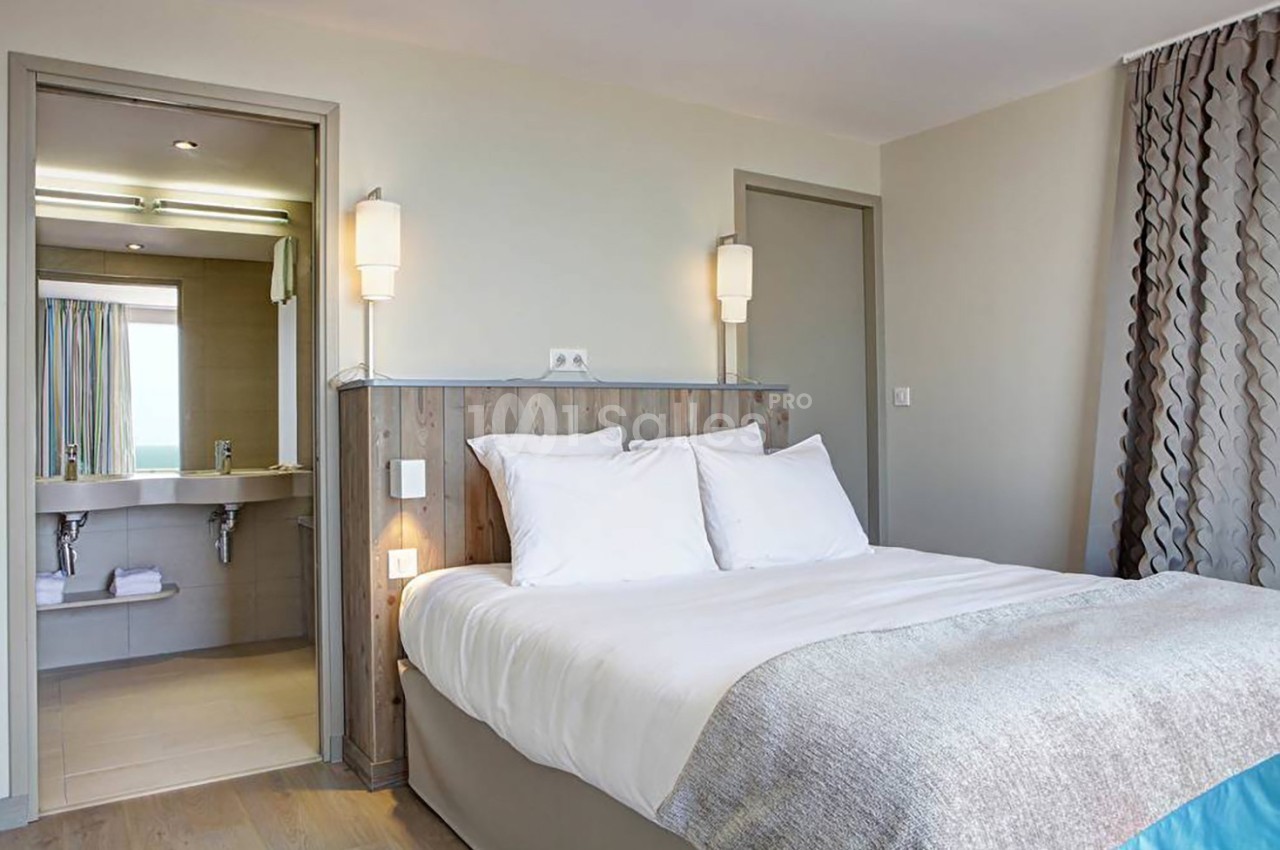 Chambre lumineuse avec lit double, tête de lit en bois, vue sur une salle de bain ouverte et rideaux à motifs.