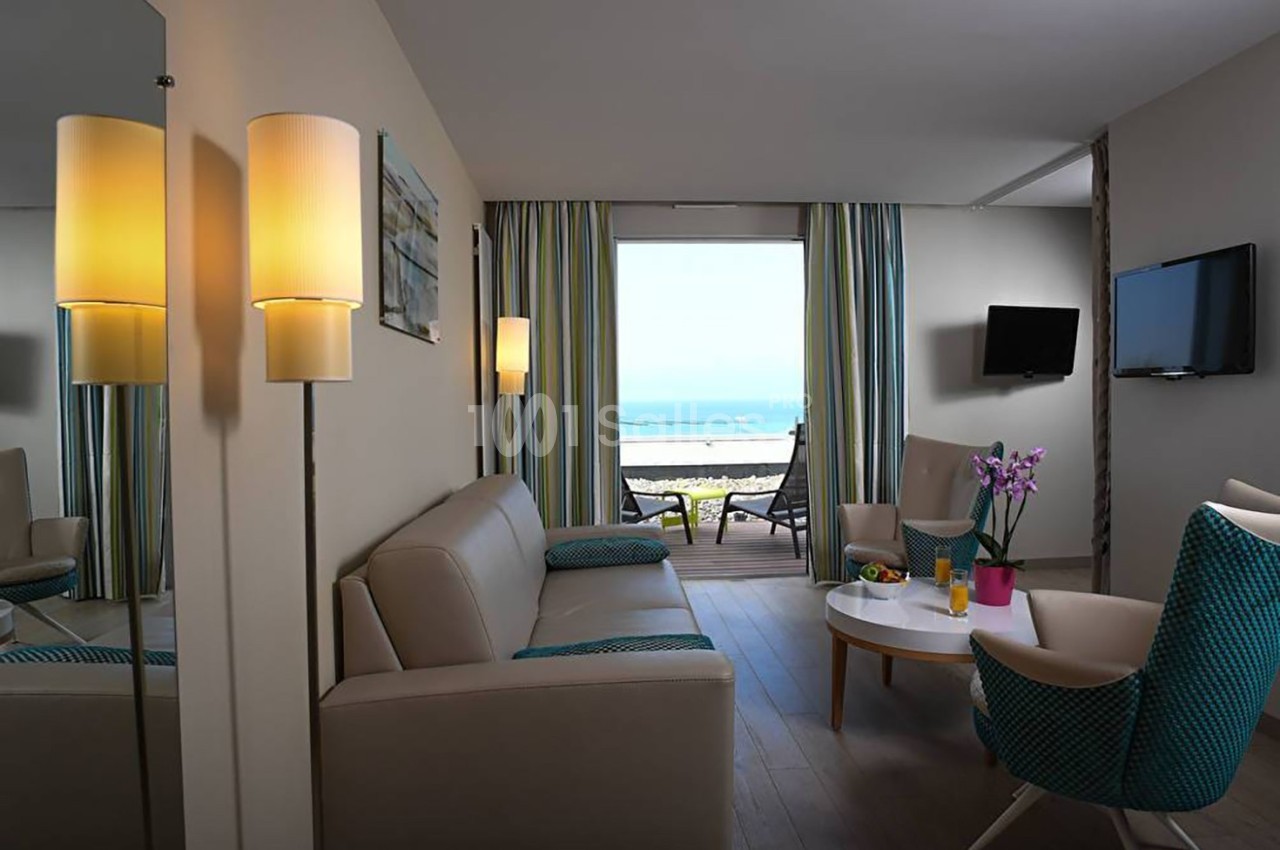 Salon lumineux avec canapé, fauteuils, table basse, téléviseur mural et vue sur la mer à travers une baie vitrée.
