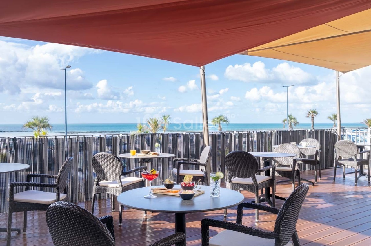 Terrasse ensoleillée avec tables et chaises, vue sur la mer, protégée par des voiles d'ombrage colorées.