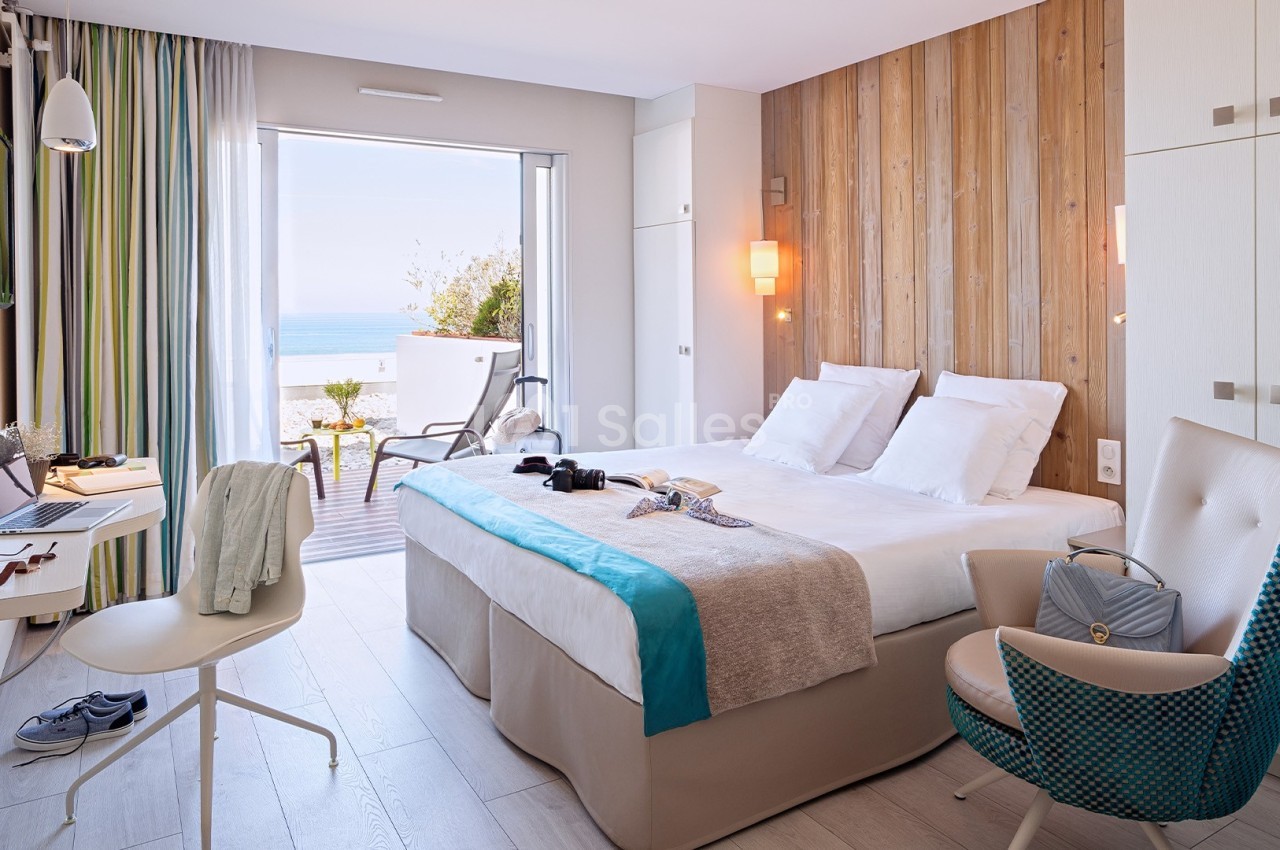 Chambre lumineuse avec lit double, bureau, fauteuil, balcon donnant sur la mer et décoration moderne en bois clair.
