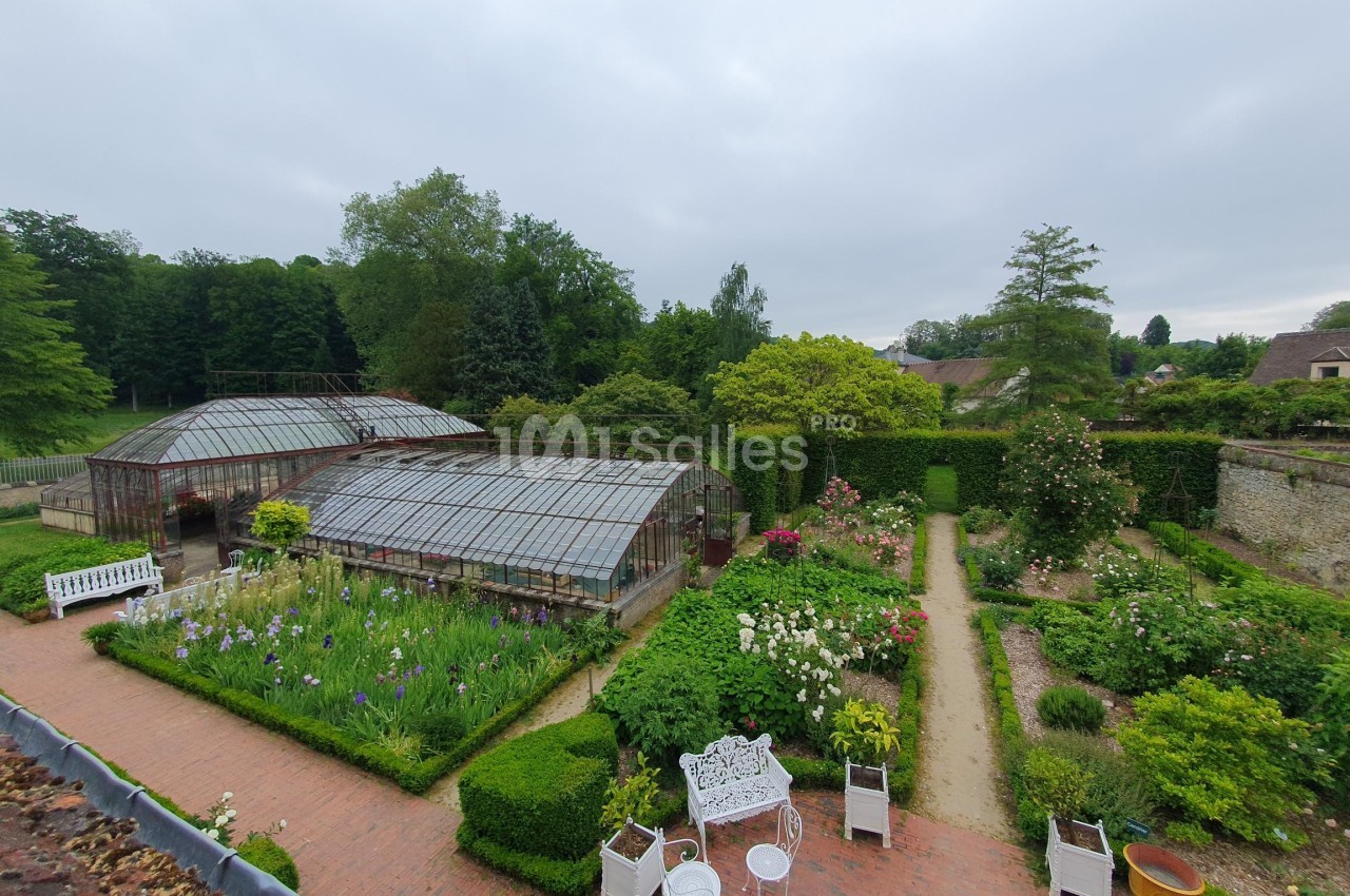 Jardin avec serres vitrées, allées bordées de fleurs, bancs blancs et végétation dense sous un ciel nuageux.