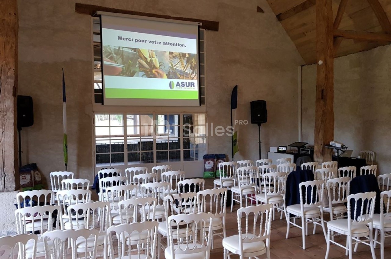 Salle de conférence avec des chaises blanches alignées, un écran de projection allumé et des équipements audio.