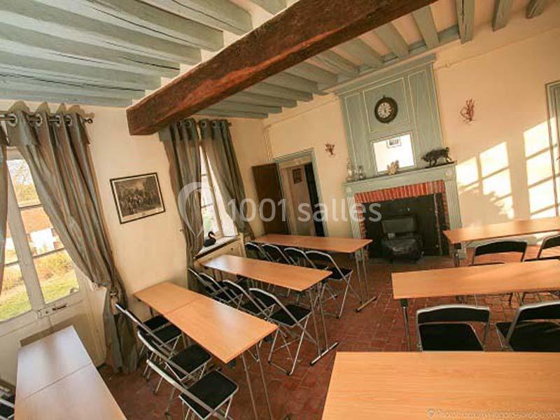 Salle lumineuse avec tables en bois, chaises pliantes, cheminée, poêle à bois et poutres apparentes au plafond.