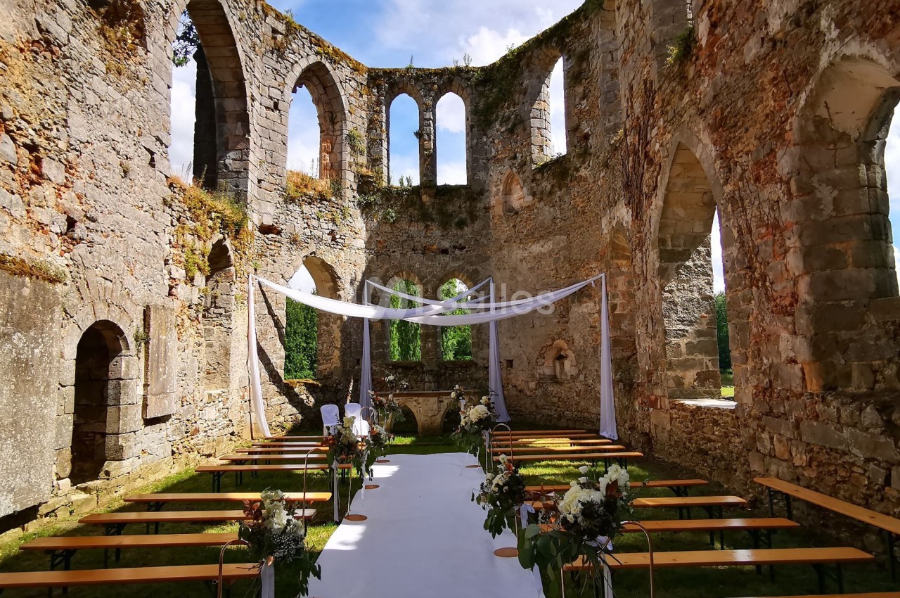 Ruines d'une église transformées en lieu de cérémonie, avec bancs alignés et une arche décorée de voilages blancs.