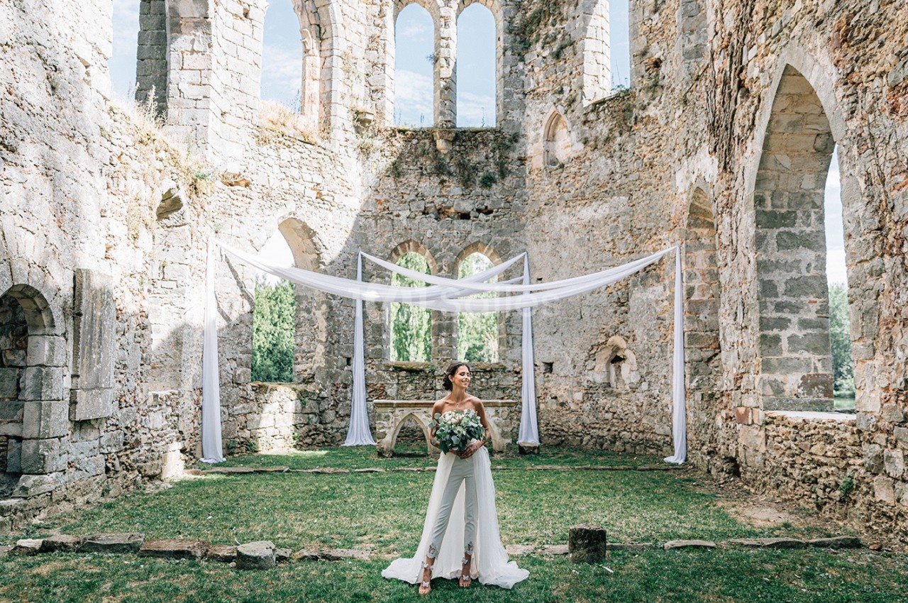 Une femme en tenue de mariage tient un bouquet dans les ruines d'une ancienne bâtisse en pierre, décorée de voilages blancs.