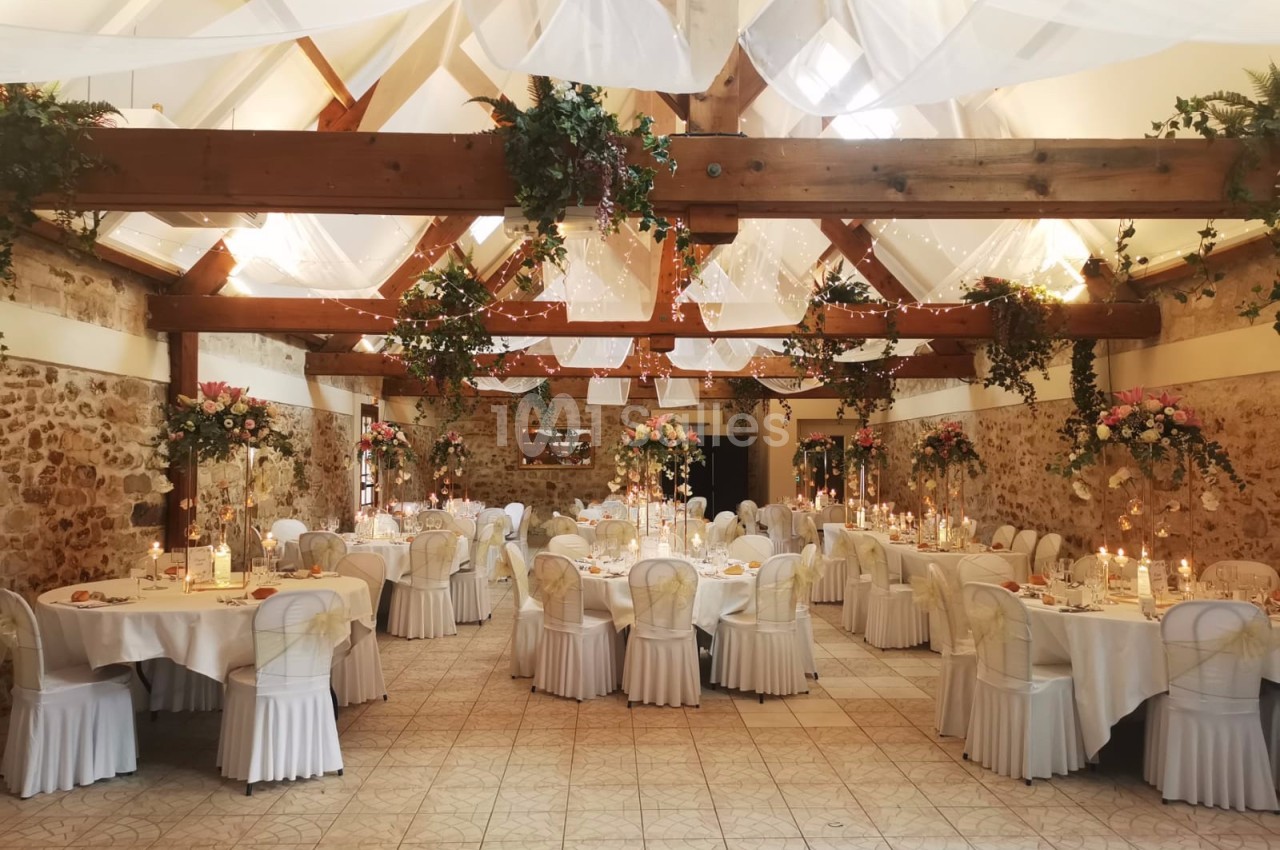 Salle de réception décorée avec des tables rondes, nappes blanches, chandeliers et guirlandes florales suspendues.